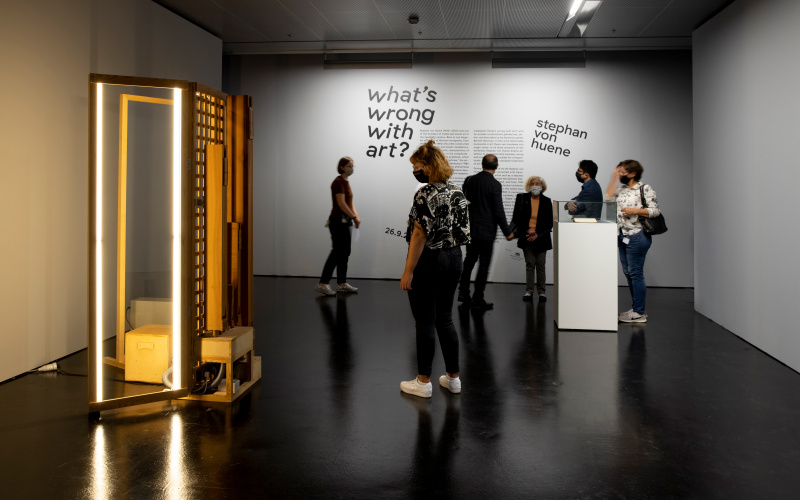 Einblick in die Ausstellung Stephan von Huene. Es sind 4 Menschen im Raum, links ist eine erleuchtete, rechteckige Standskulptur